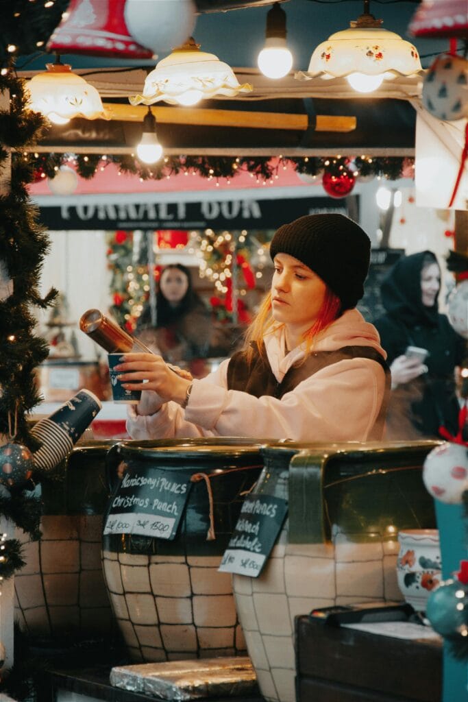 Vendedora preparando bebida quente em mercado de Natal na Europa com decoração festiva.