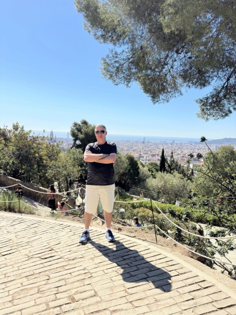 Homem posando em pé no Parque Güell, com a cidade de Barcelona ao fundo sob céu azul e ensolarado.
