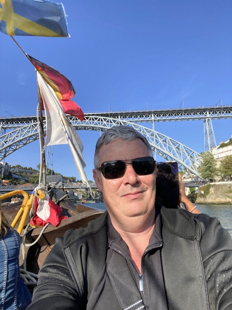 Selfie de um homem em um barco no Rio Douro, com a Ponte Dom Luís I ao fundo em um dia ensolarado em Porto, Portugal.
