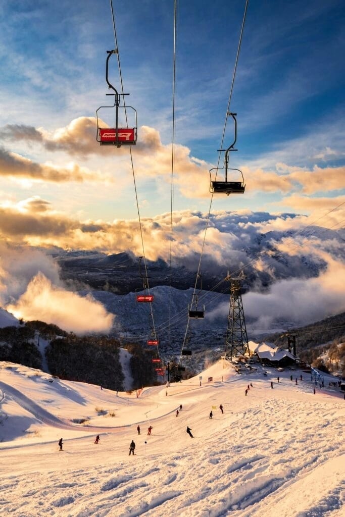 Pessoas esquiando no Cerro Catedral em Bariloche ao entardecer, com teleférico e céu alaranjado.