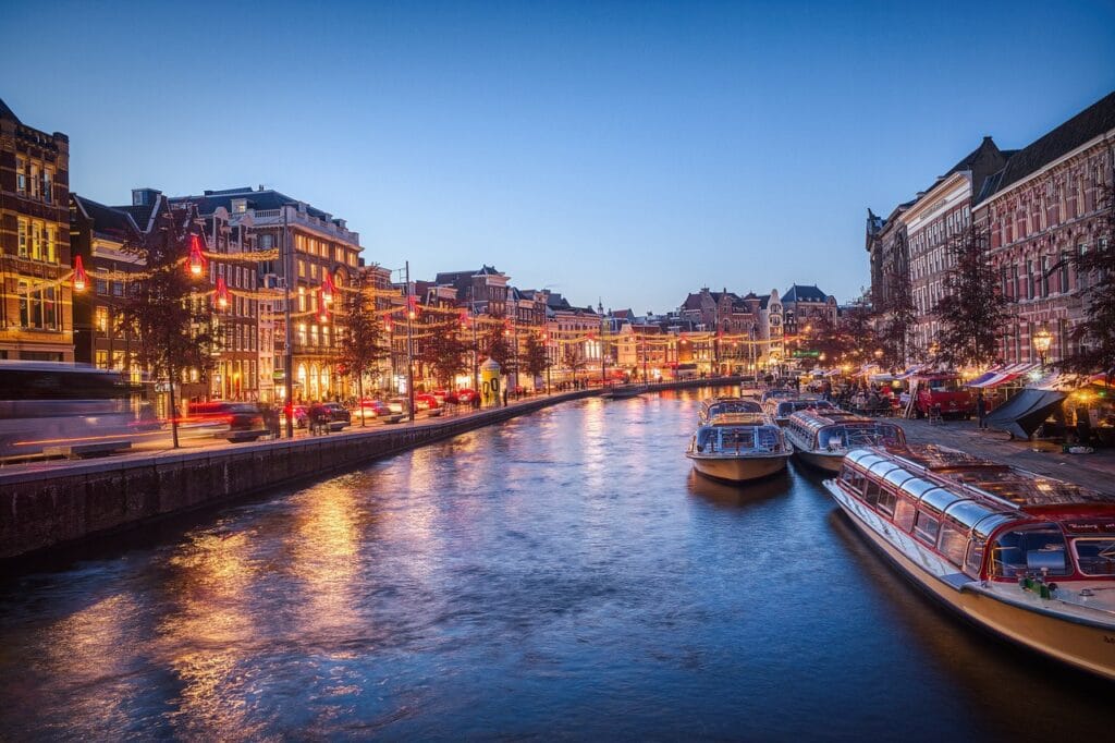Canal de Amsterdã ao entardecer com barcos turísticos e iluminação urbana.