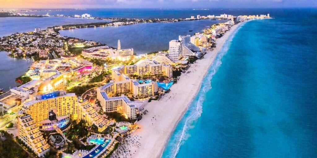 Vista aérea de Cancún com resorts iluminados, praia de areia branca e mar azul-turquesa.