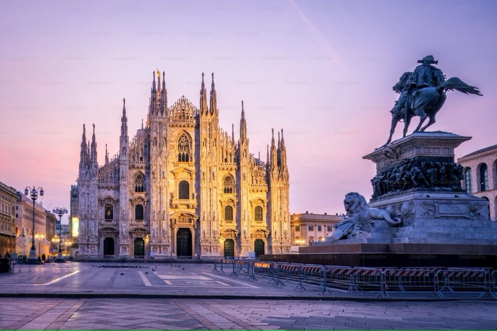Fachada da Catedral de Milão (Duomo) iluminada ao amanhecer, com a estátua equestre de Vittorio Emanuele II em primeiro plano.