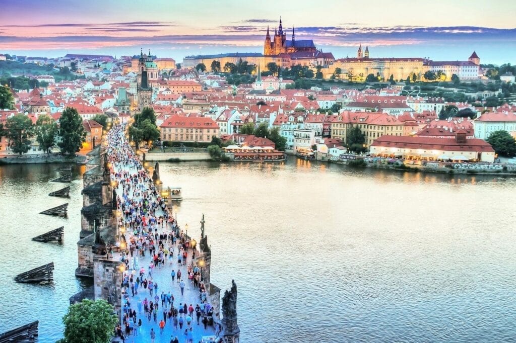 Ponte Carlos em Praga repleta de turistas com vista para o castelo iluminado ao fundo.