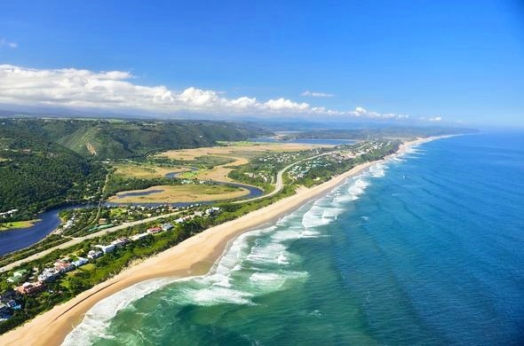 Vista aérea da praia de Wilderness com mar azul, montanhas verdes e casas costeiras na África do Sul.