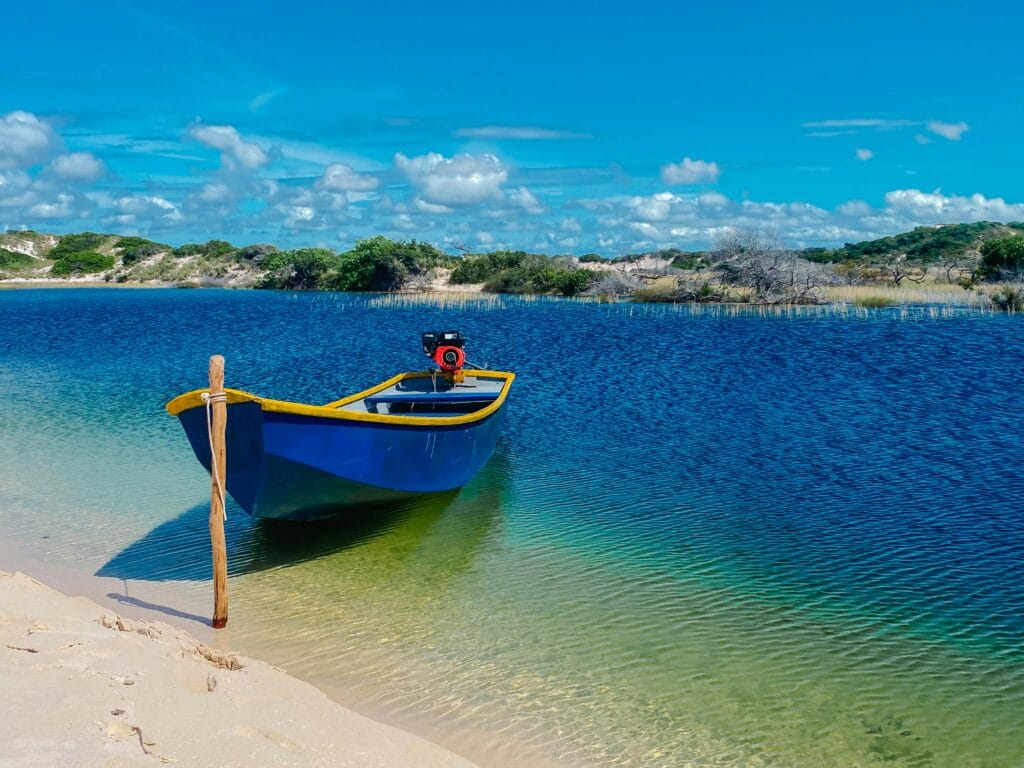 Barco azul ancorado em lagoa de águas cristalinas nos Lençóis Maranhenses, cercada por vegetação. Planeje e curta os Lençóis Maranhenses sem erro!