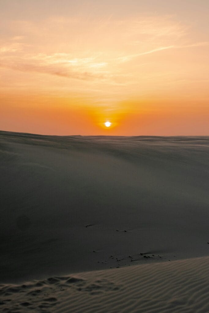 Pôr do sol visto entre as dunas dos Lençóis Maranhenses, com céu alaranjado e relevo ondulado de areia