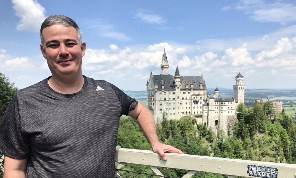 Blogueiro posando em frente ao Castelo de Neuschwanstein na Alemanha, com o castelo ao fundo em um dia ensolarado. Bate e volta ao Castelo de Neuschwanstein.