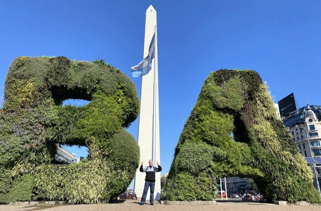 Carlos posando entre as letras "BA" feitas de plantas, com o Obelisco ao fundo em Buenos Aires. Roteiro de 6 dias em Buenos Aires.