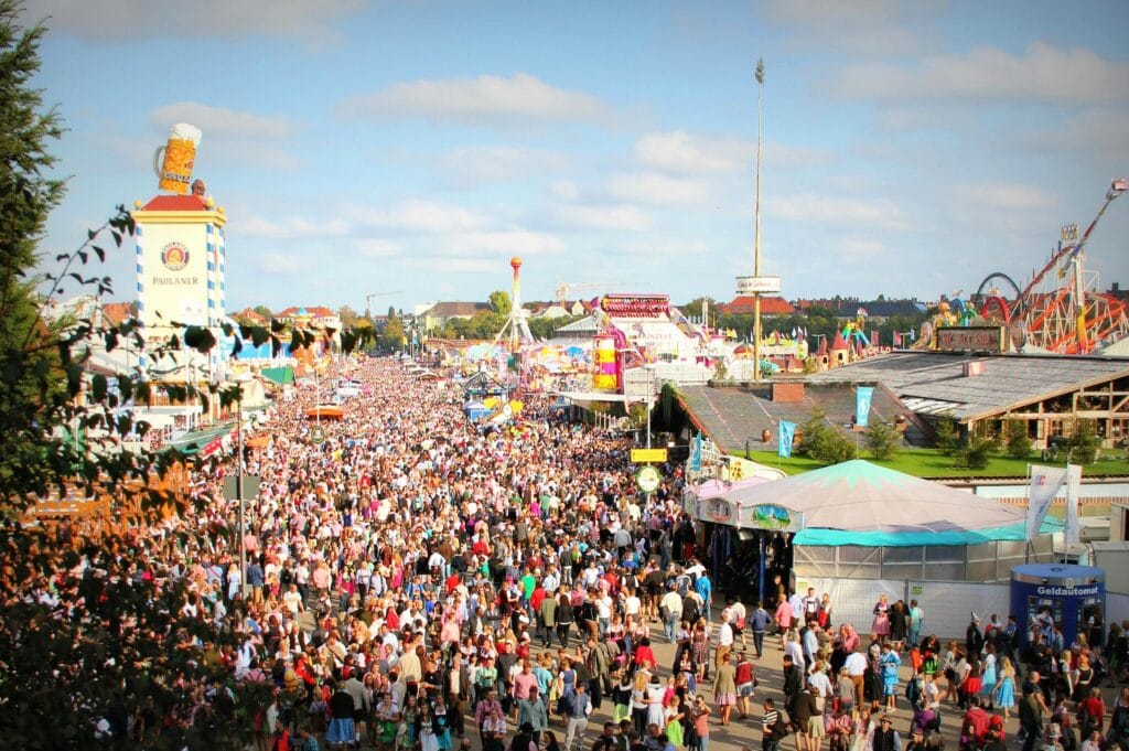 Multidão animada vestindo trajes típicos durante a Oktoberfest em Munique, com barracas de cerveja e atrações de parque ao fundo. Roteiro de 5 dias em Munique.